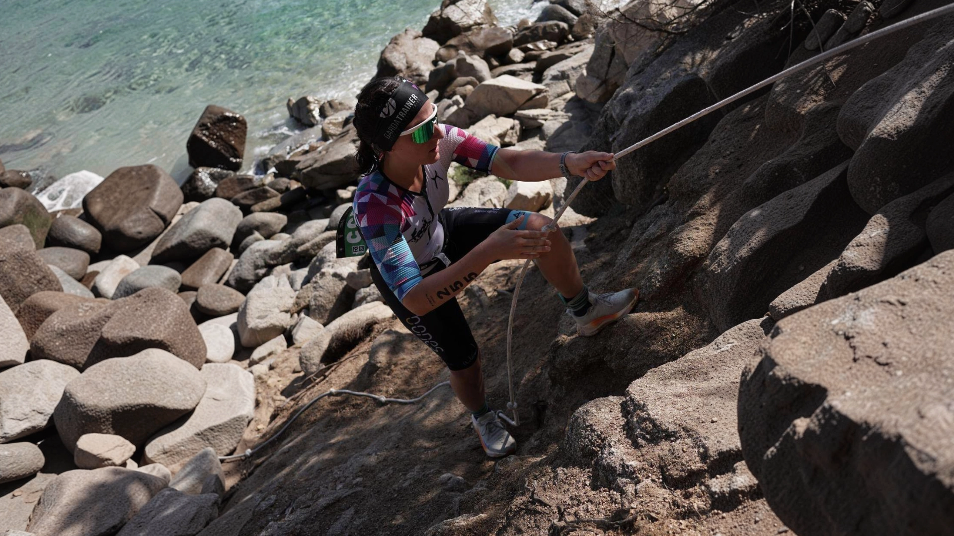 Atleta in divisa From Zero to Tri risale un tratto roccioso sulla costa aiutandosi con una corda.