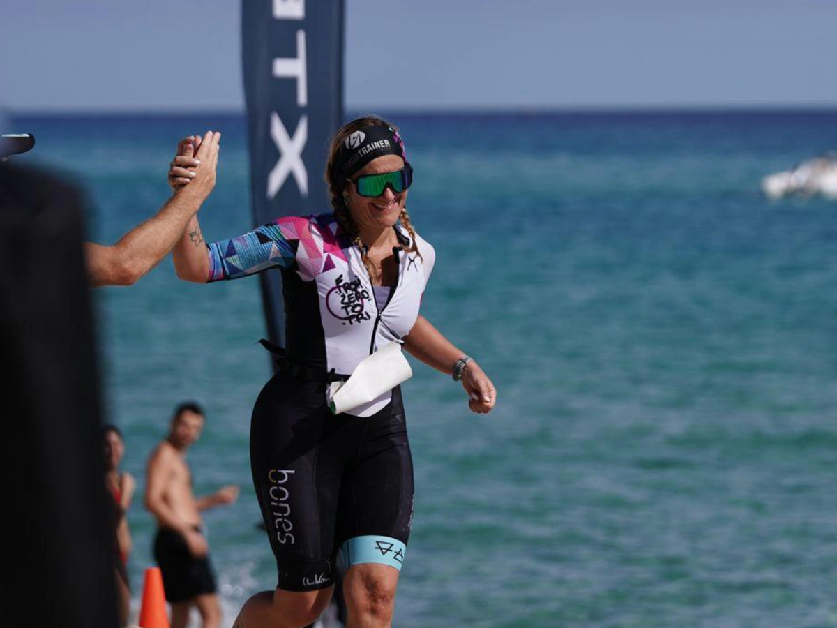 Atleta in divisa From Zero to Tri sorride e dà il cinque al traguardo, con il mare sullo sfondo.
