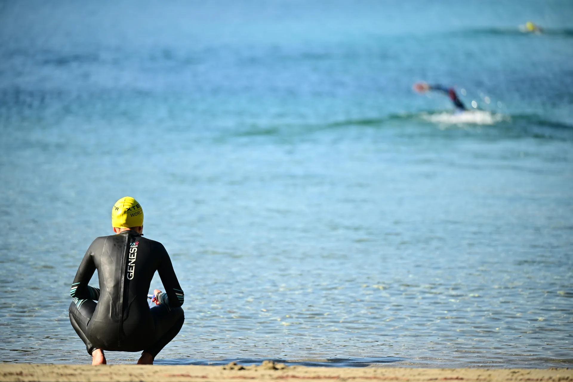 Atleta in muta e cuffia gialla accovacciato sulla riva, rivolto verso il mare.