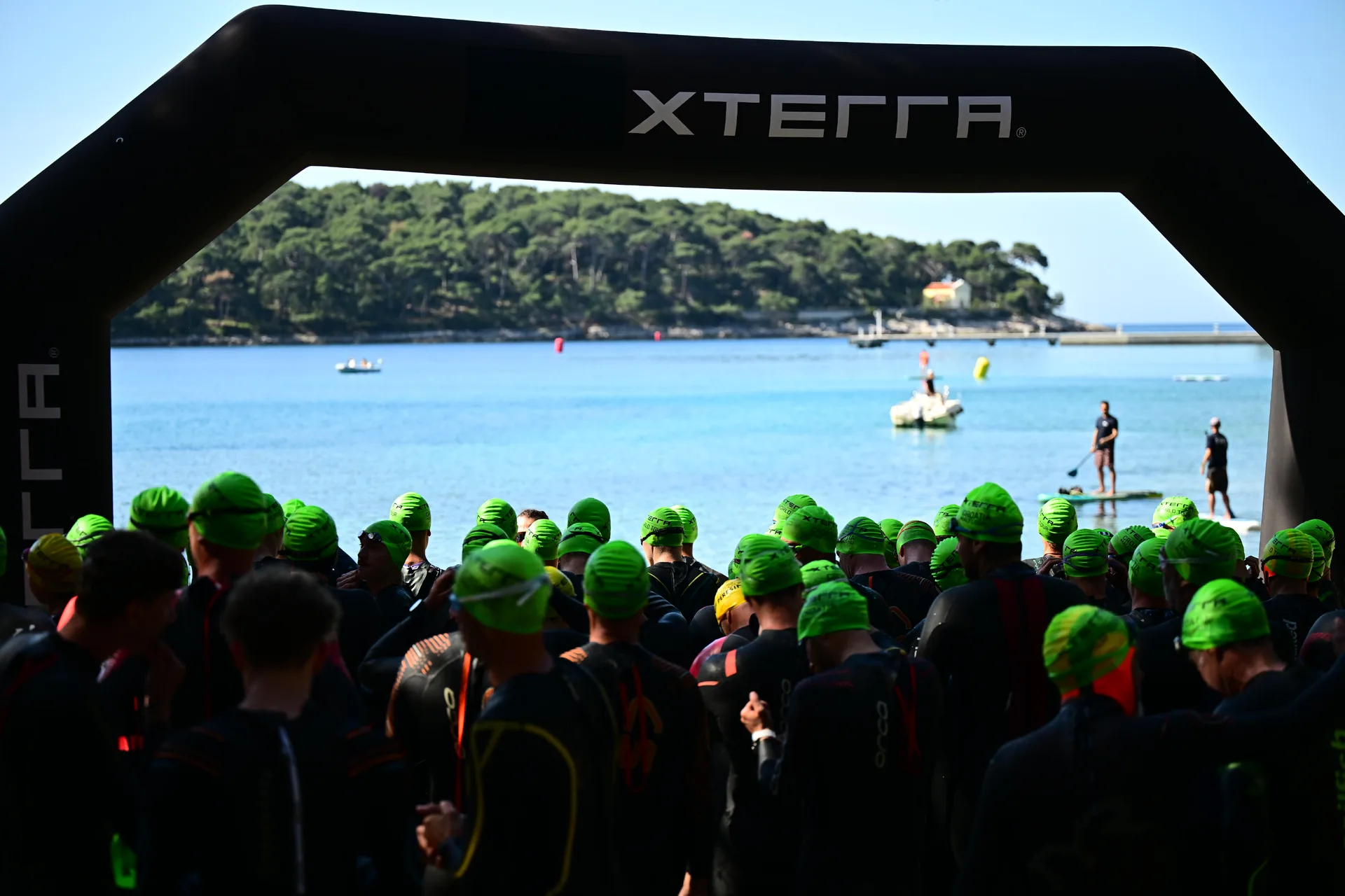 Atleti in muta e cuffia verde sotto l’arco XTERRA, rivolti verso la baia.