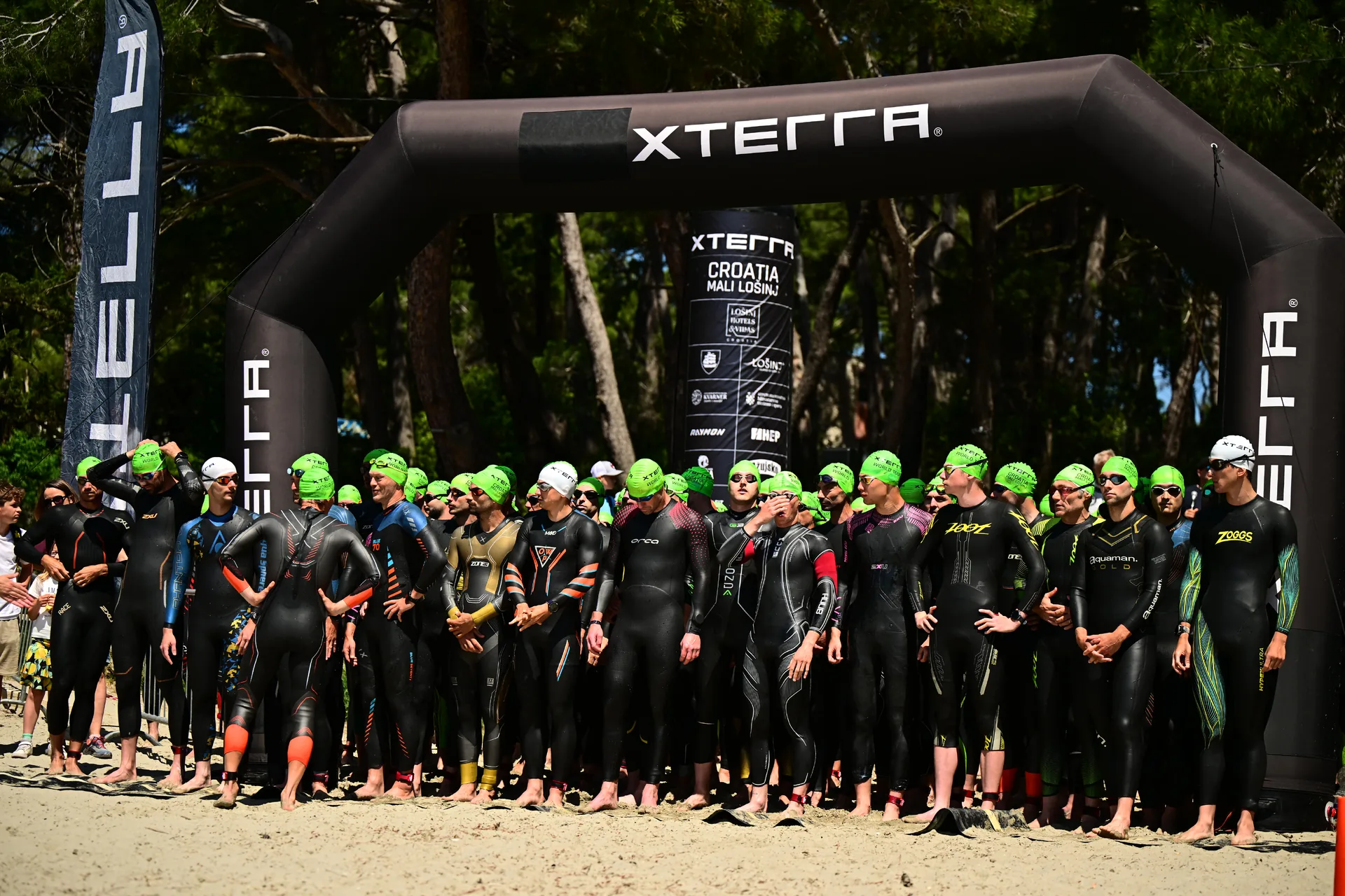 Atleti in muta e cuffia verde schierati sulla spiaggia sotto l’arco XTERRA.